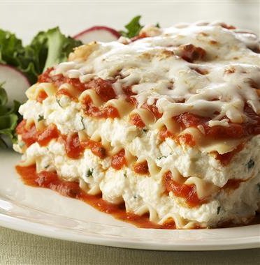 A piece of Ricotta Cheese Lasagna on a white plate. A small salad is on the plate behind the lasagna.