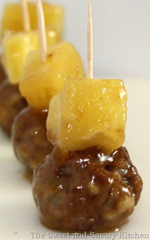 A row of Aloha Meatballs on a white dish. Each meatball topped with pineapple., and has a wooden toothpick going through it for servings.